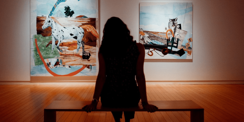 A woman sitting on top of a wooden bench in front of two paintings.