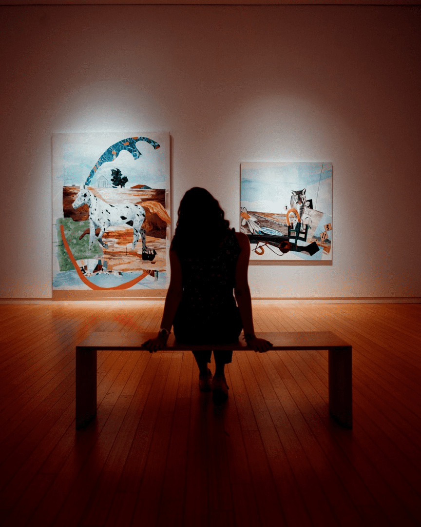A woman sitting on top of a wooden bench in front of two paintings.